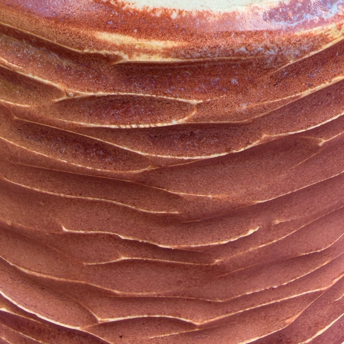 A handmade ceramic bottle with a textured, ridged brown body and a glossy white neck, displayed on a reddish-brown surface.