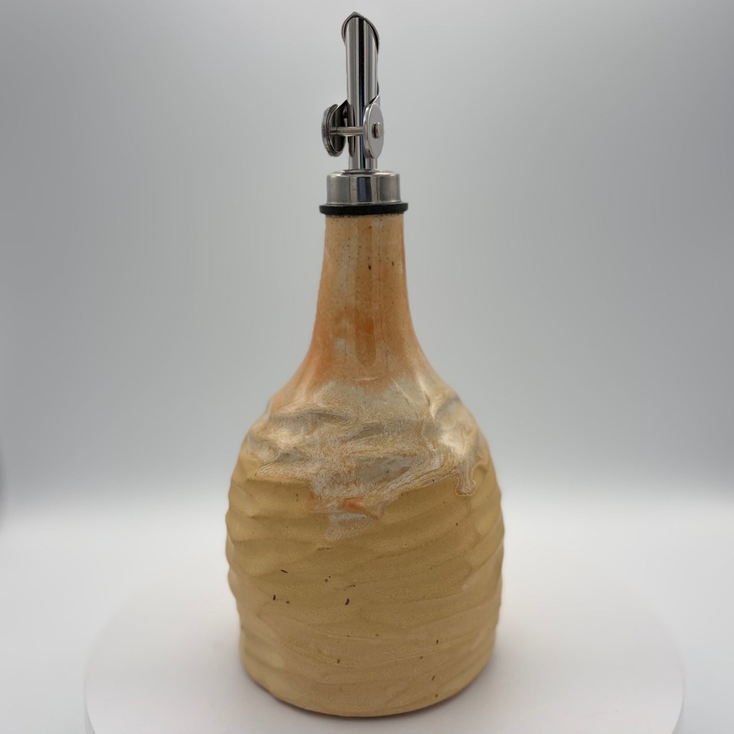 A handmade ceramic bottle with a textured, ribbed surface in a warm beige and light brown glaze, featuring a silver metal pour spout with a black rubber collar, set against a neutral gray background.