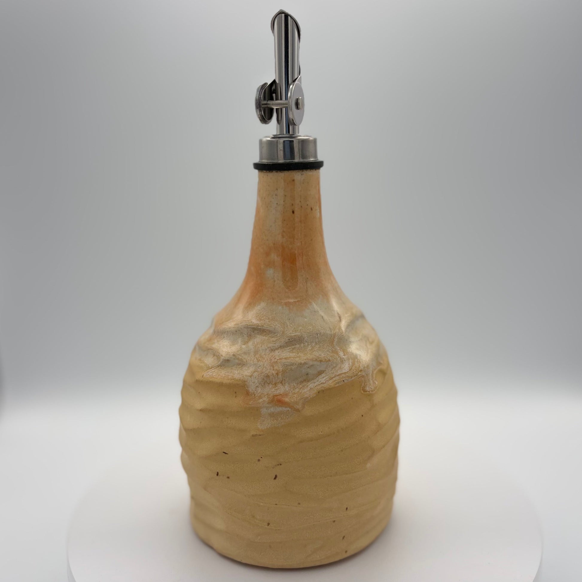 A handmade ceramic bottle with a textured, ribbed surface in a warm beige and light brown glaze, featuring a silver metal pour spout with a black rubber collar, set against a neutral gray background.