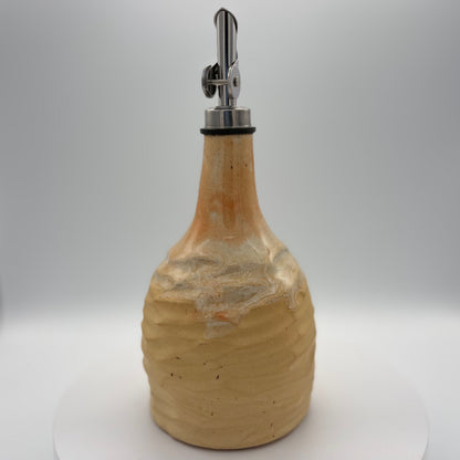 A handmade ceramic bottle with a textured, ribbed surface in a warm beige and light brown glaze, featuring a silver metal pour spout with a black rubber collar, set against a neutral gray background.