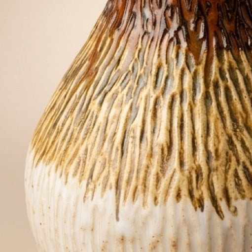 A handmade ceramic bottle with a cork stopper, featuring a textured glaze that transitions from a warm cream-white at the bottom to a dark brown and golden-brown at the top, set against a solid beige background.