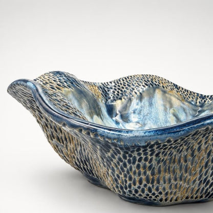 A decorative ceramic bowl with a wavy, organic silhouette, featuring a textured blue and gold patterned glaze on a light gray background.