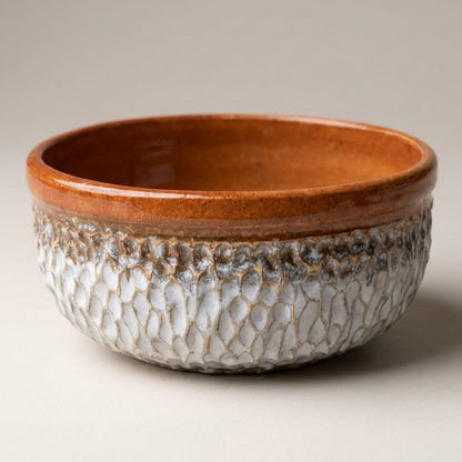 A handmade ceramic bowl with a textured, scale-like glazed pattern in off-white and brown, featuring a smooth, warm brown rim, set against a neutral light gray background.