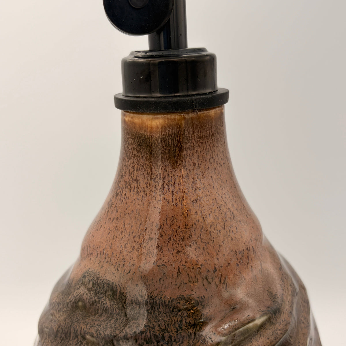 A close-up of a ceramic pump bottle with a textured, speckled brown and black glaze, featuring a black plastic pump dispenser on a light gray background.
