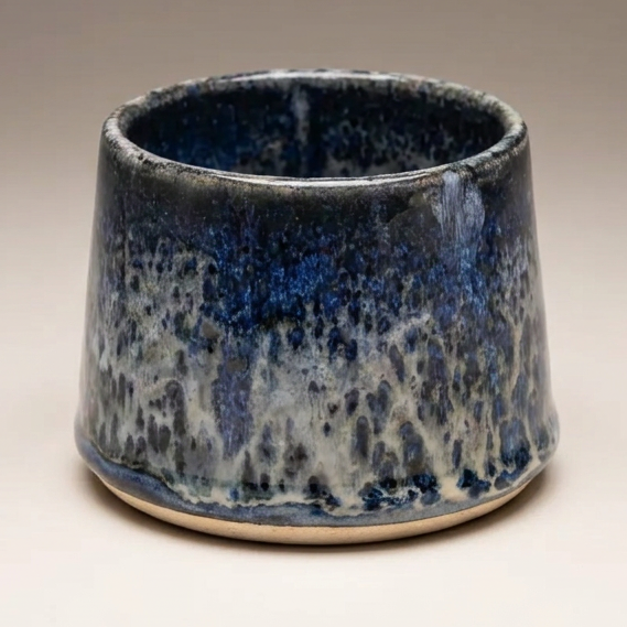 A small ceramic cup with a tapered shape, featuring a dark blue and white mottled glaze, set against a neutral gradient background.