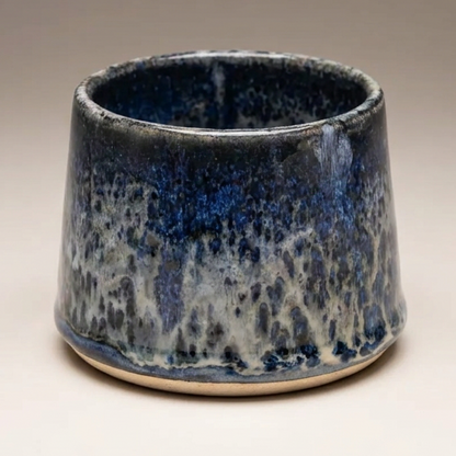 A small ceramic cup with a tapered shape, featuring a dark blue and white mottled glaze, set against a neutral gradient background.