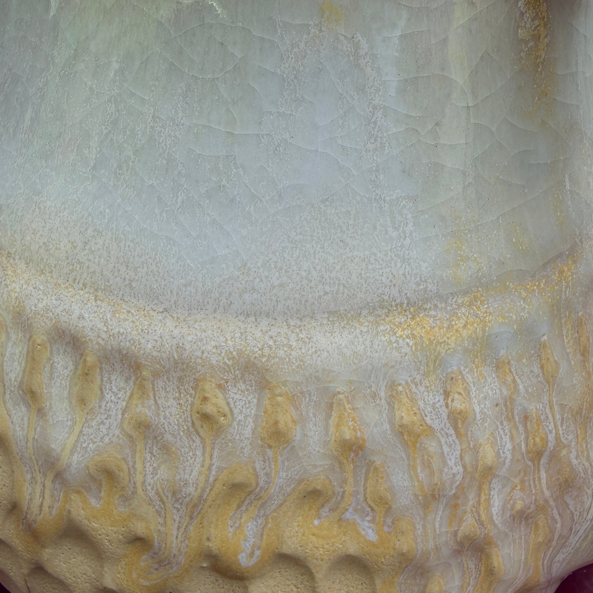 Close-up of a ceramic pitcher or vase with a textured, bubbled lower half and a smooth upper body, featuring a cream to pale yellow glaze with golden-yellow dripped accents.