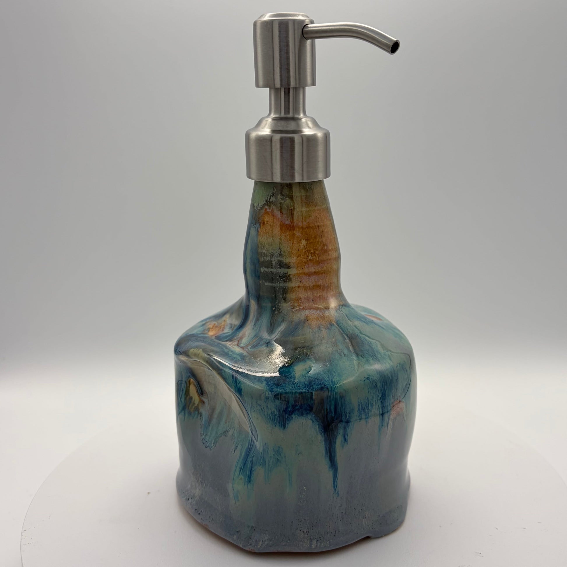 A handmade ceramic soap dispenser with a textured, organic shape in blue, teal, and brown glazes, topped with a brushed silver stainless steel pump, shown on a white circular platform against a light gray background.