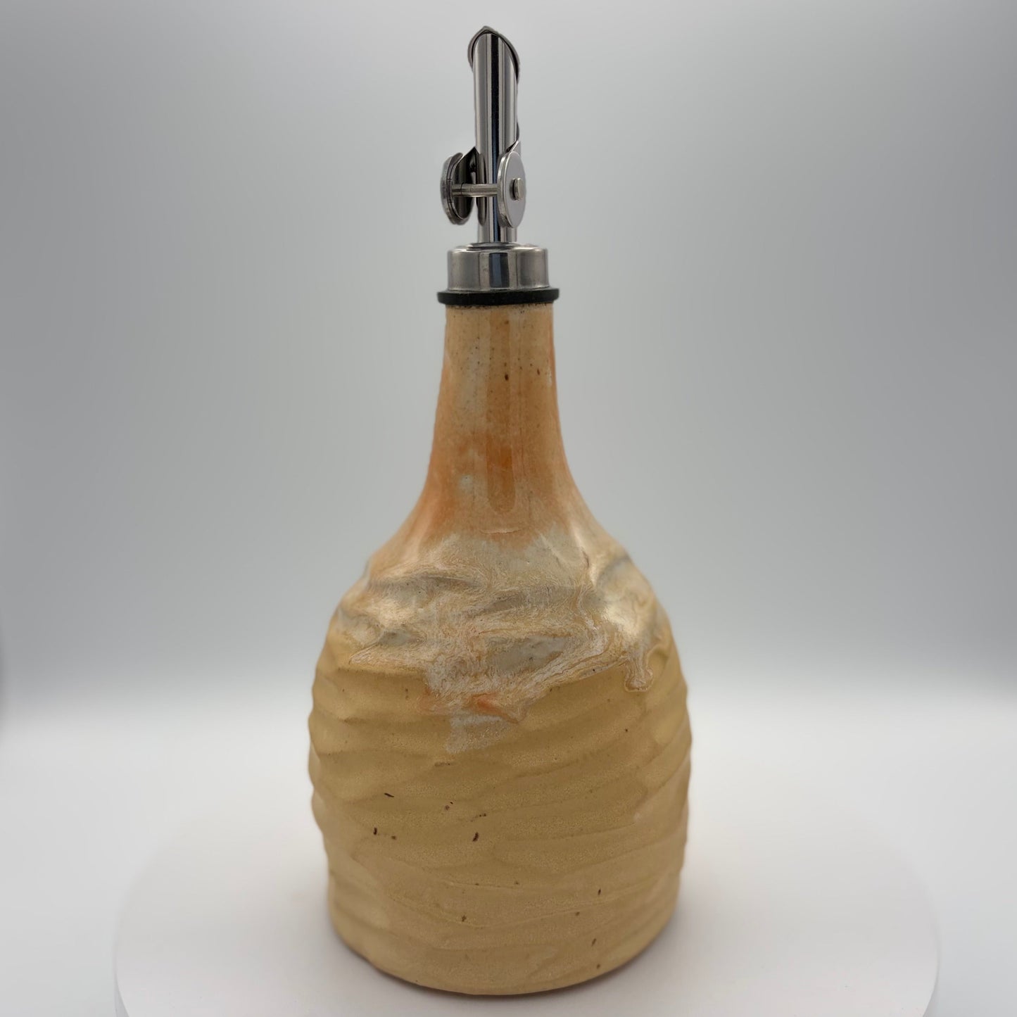 A handmade ceramic bottle with a textured, ribbed surface in a warm beige and light brown glaze, featuring a silver metal pour spout with a black rubber collar, set against a neutral gray background.