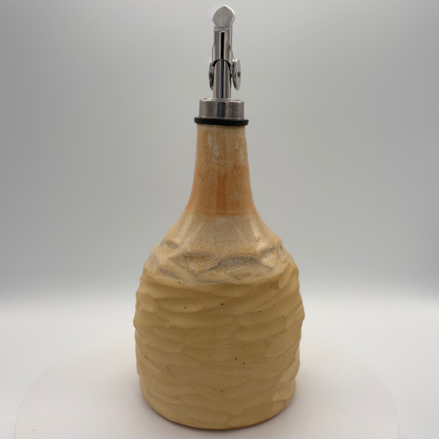 A handmade ceramic oil bottle with a textured, ribbed body in a warm beige stoneware finish, featuring a polished silver metal pour spout with a black rubber gasket, set against a neutral white studio background.