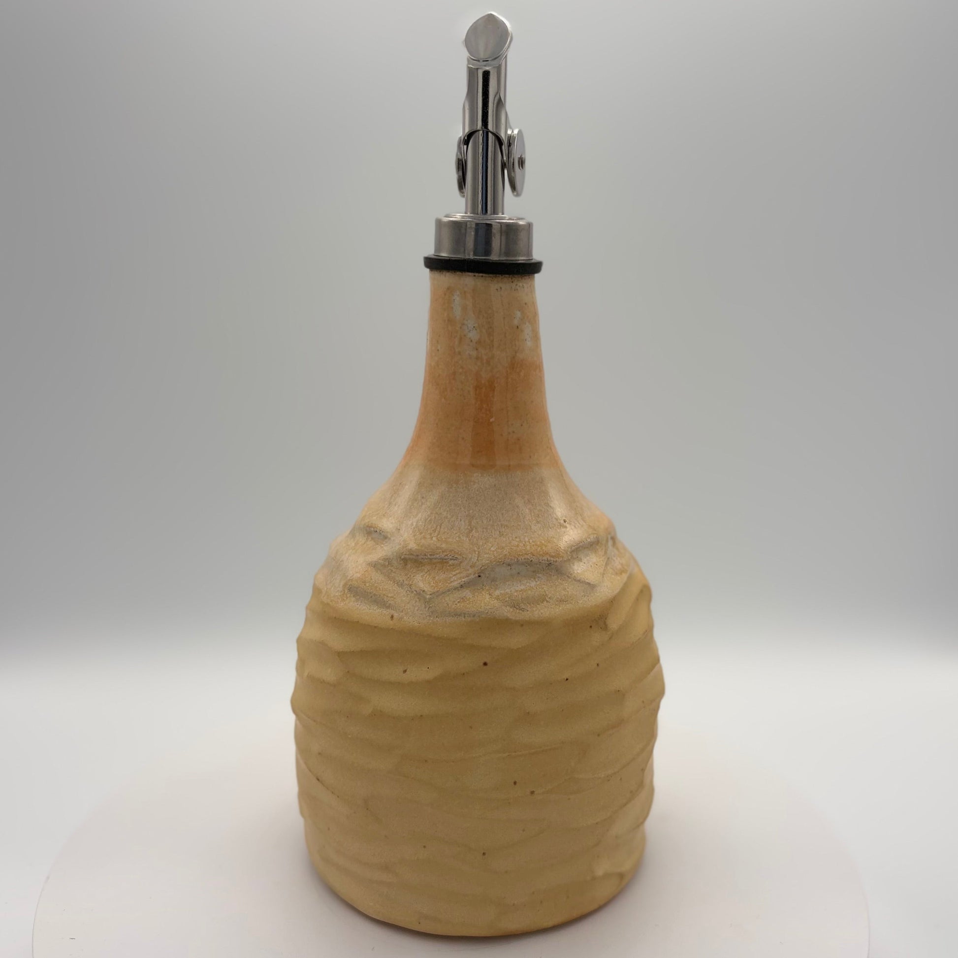 A handmade ceramic oil bottle with a textured, ribbed body in a warm beige stoneware finish, featuring a polished silver metal pour spout with a black rubber gasket, set against a neutral white studio background.