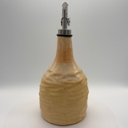 A handmade ceramic oil bottle with a textured, ribbed body in a warm beige stoneware finish, featuring a polished silver metal pour spout with a black rubber gasket, set against a neutral white studio background.