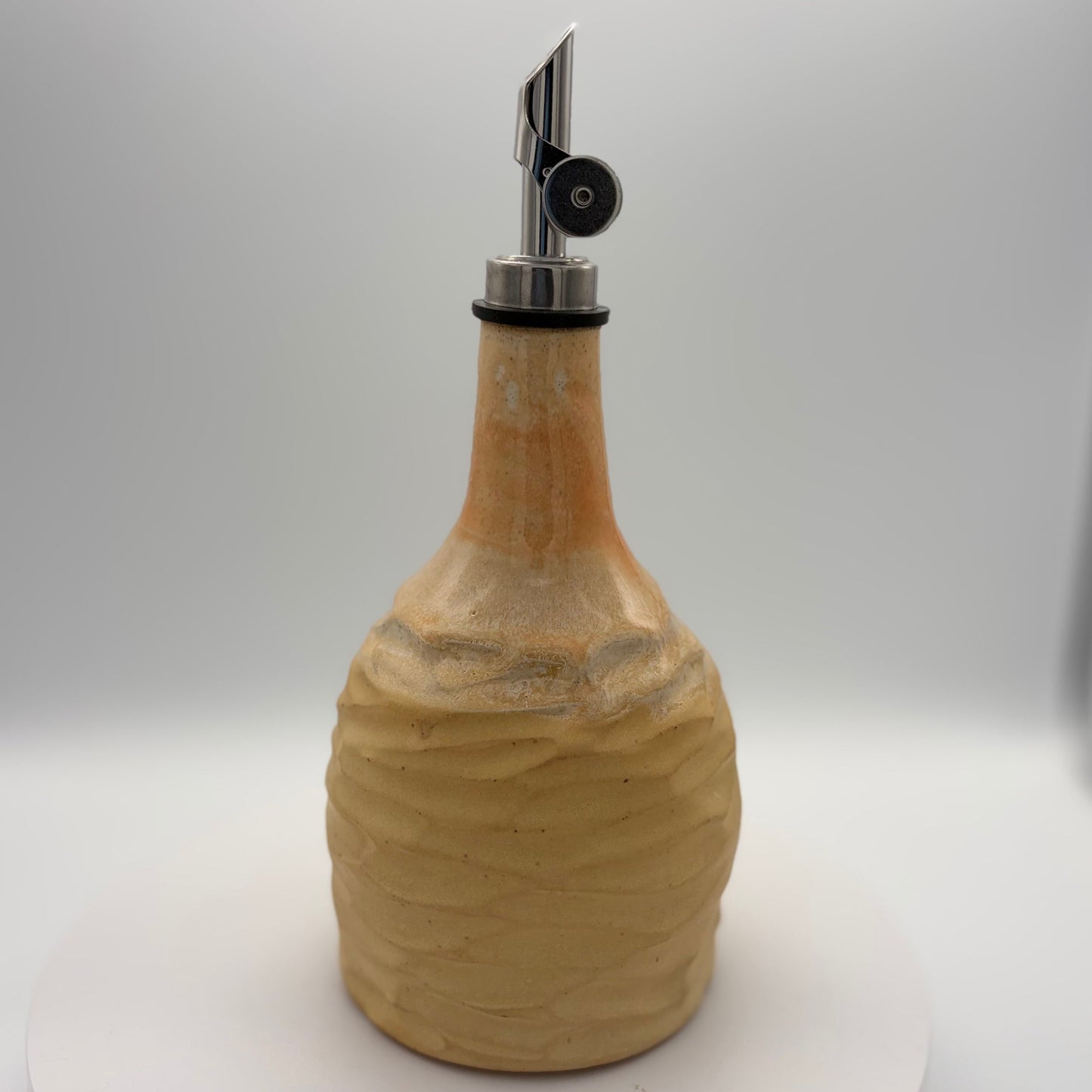 A ceramic oil dispenser bottle with a textured, ribbed beige body and a polished silver metal pour spout, set against a neutral gray studio background.