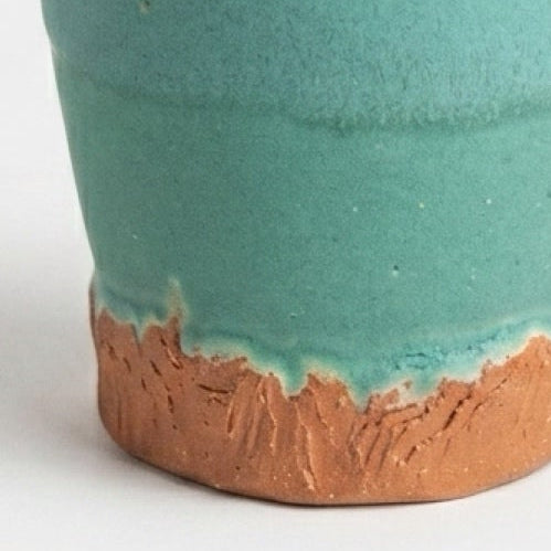 A handmade ceramic cup with a textured teal-green glaze and a rustic terracotta-colored unglazed base, shown on a light gray background.