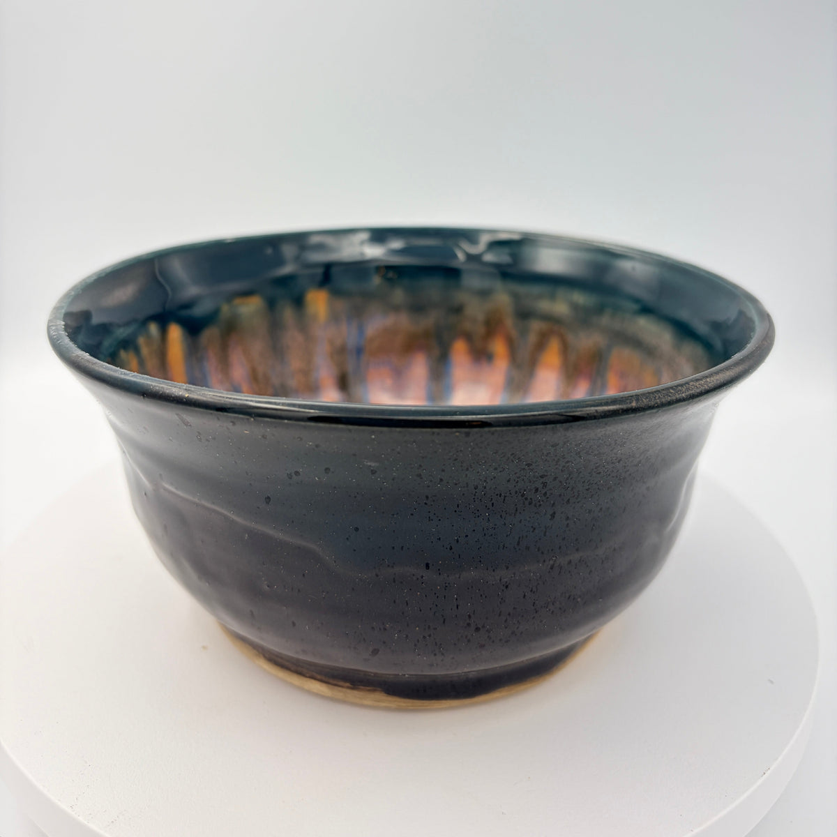 A handmade ceramic bowl with a glossy black and dark blue glaze, featuring a warm orange and yellow iridescent crackle pattern inside, displayed on a white circular riser against a white studio background.