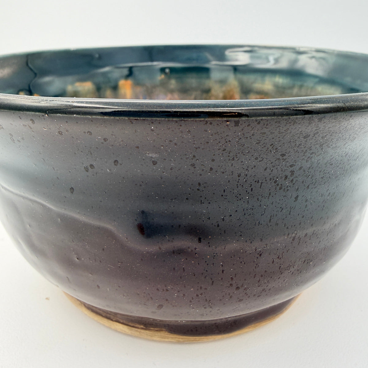 A close-up of a handmade ceramic bowl with a dark, speckled glaze that transitions from deep black to a subtle blue-green at the rim, set against a plain white background.