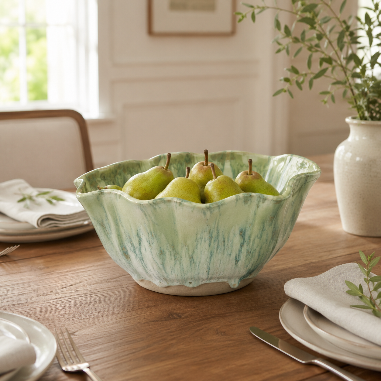 A large scalloped ceramic bowl with a light green and white marbled pattern, filled with green pears, set on a wooden dining table in a bright, neutral room.
